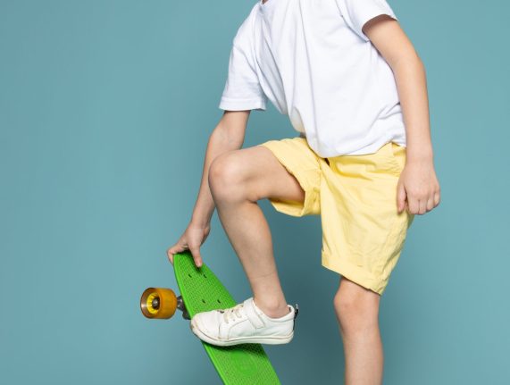 front-view-smiling-cute-kid-riding-green-skateboard-white-t-shirt-orange-shorts-blue-space